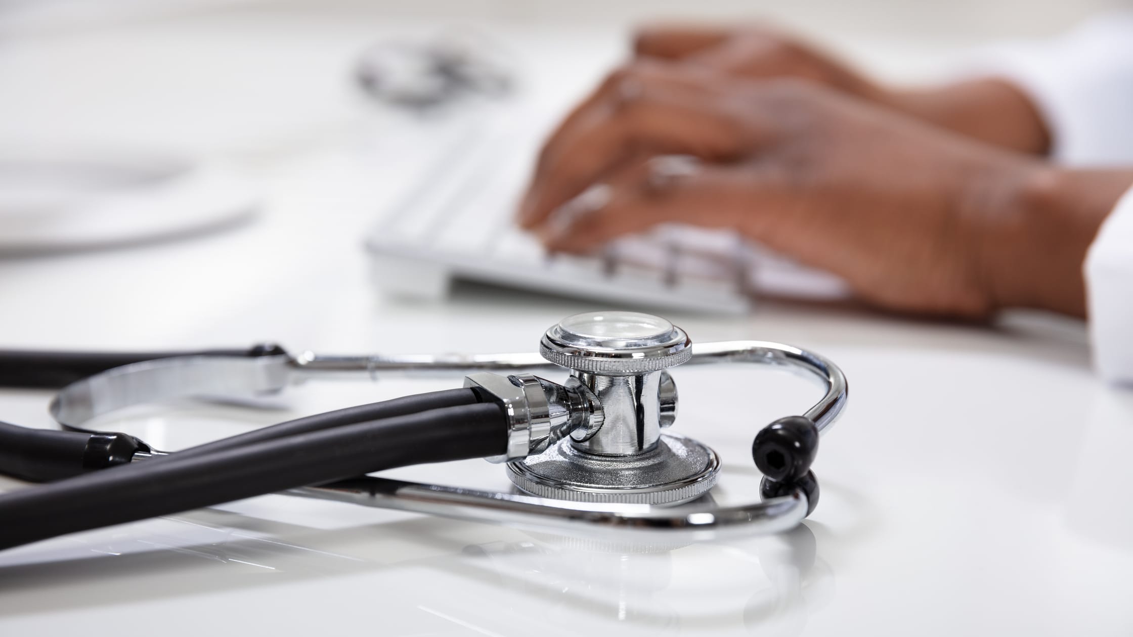 Close up image of a stethoscope with a doctor's hand typing on a keyboard in the background